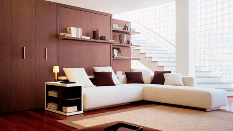 Cama abatible de matrimonio con sofá rinconero y almacenaje inferior integrado.