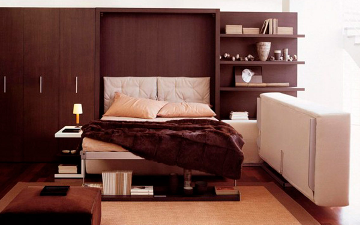 Cama abatible de matrimonio con sofá rinconero y almacenaje inferior integrado.